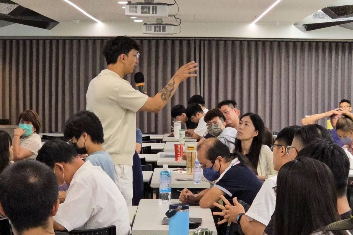 蟬師拿著麥克風站在學員座位之間，一邊比手勢一邊講解，周圍學員抬頭聽