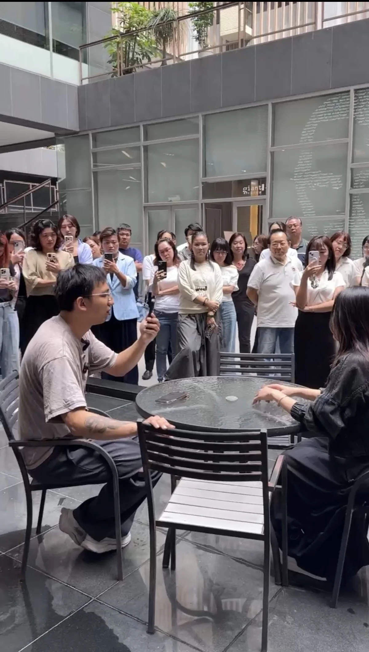中庭現場，蟬師拿著麥克風和一位學員面對面坐著示範訪談，一整圈學員圍著用手機錄影