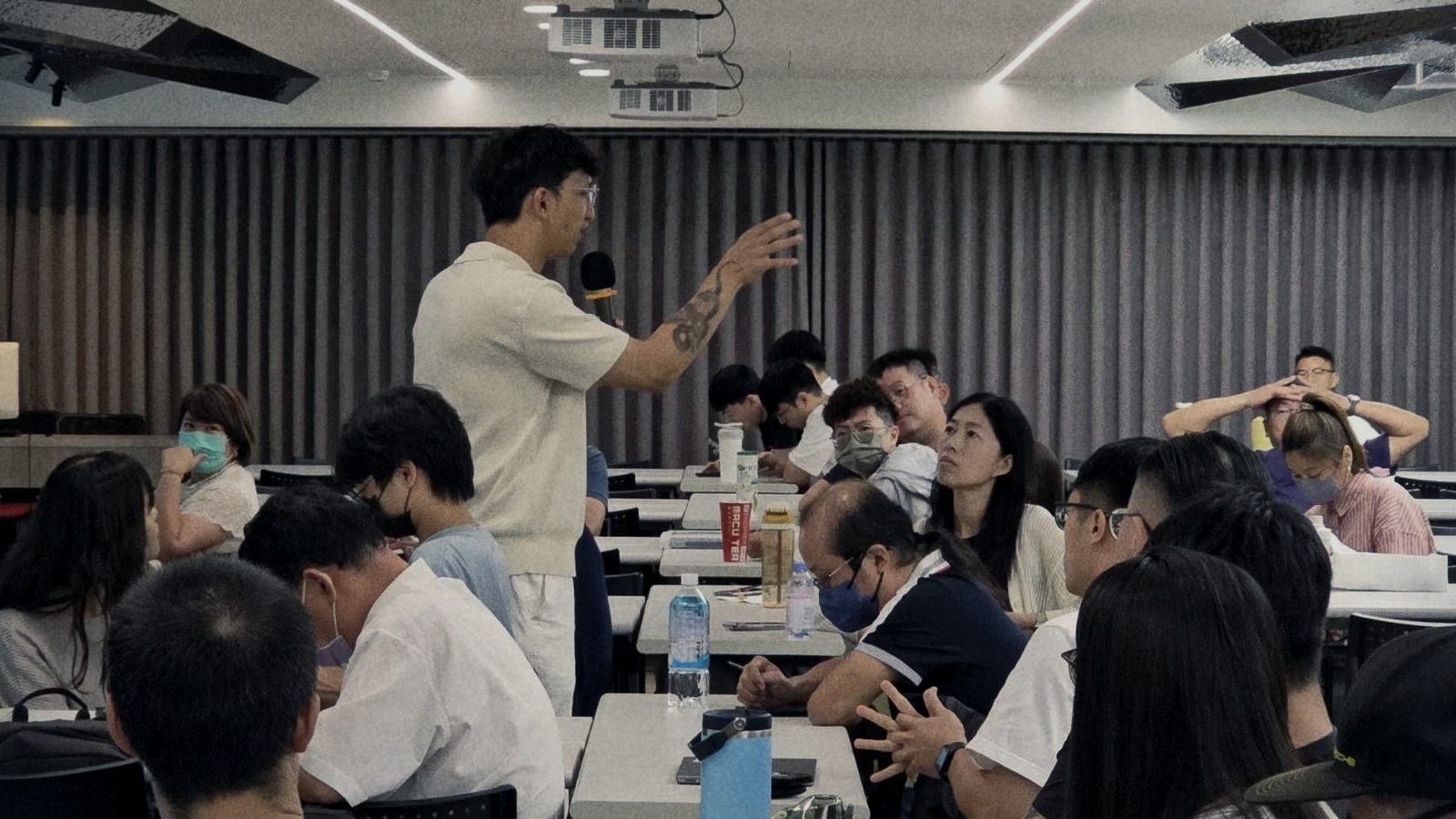 蟬師拿麥克風站在學員之間講課，手勢比向前方